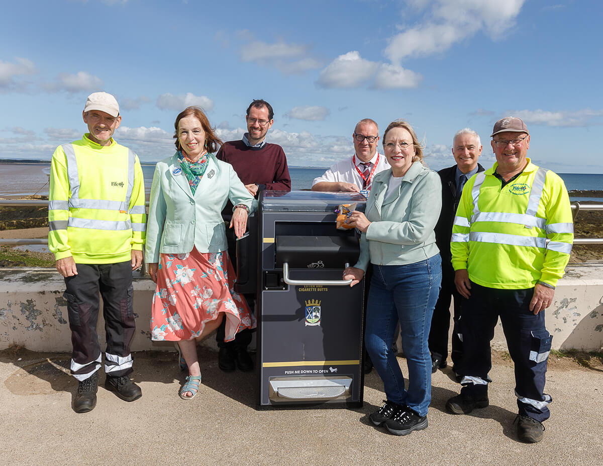 7 people standing around a litter bin with water and blue sky in the background