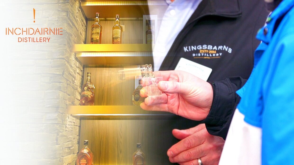 Image showing whisky bottles displayed on wooden shelves at InchDairnie Distillery, with two people in the foreground holding a small tasting glass