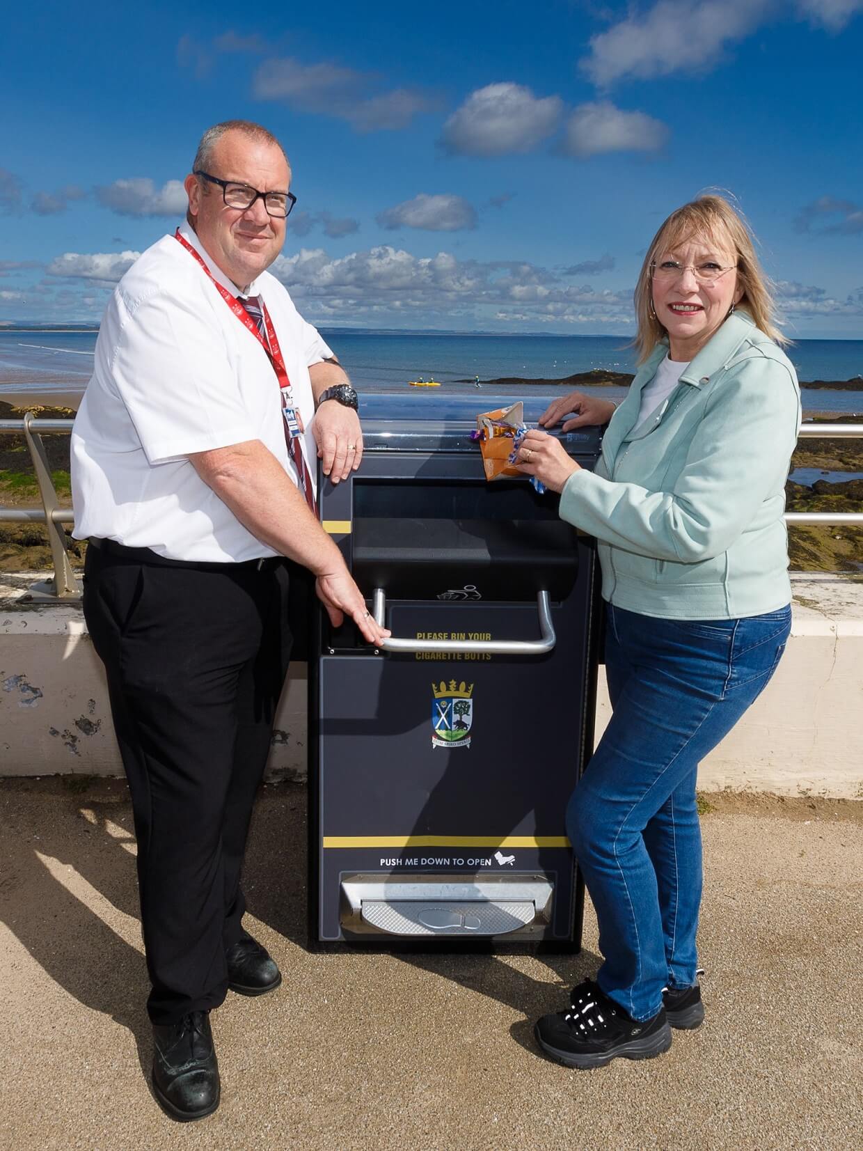 two people standing next to a litter bin holding to its handle with water a blue sky in the background