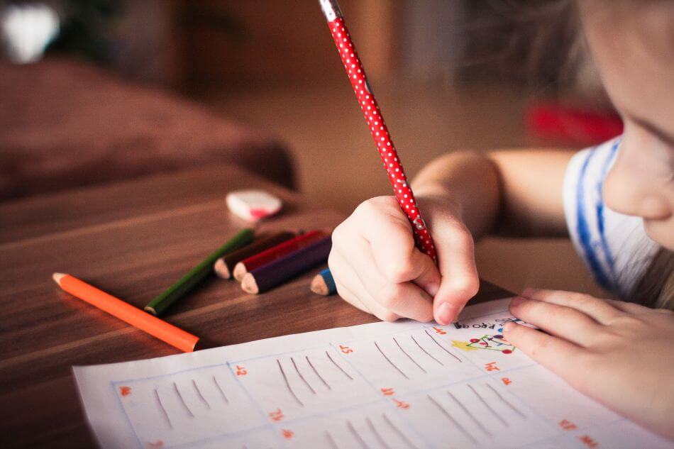 A young child in a striped t-shirt sits at a desk, writing, with coloured pencils beside them