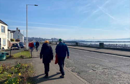 People walking along a sunny waterfront pavement beside houses and a road, with water, railings, and distant bridges visible in the background. - Community Wellbeing Project to continue in South and West Fife