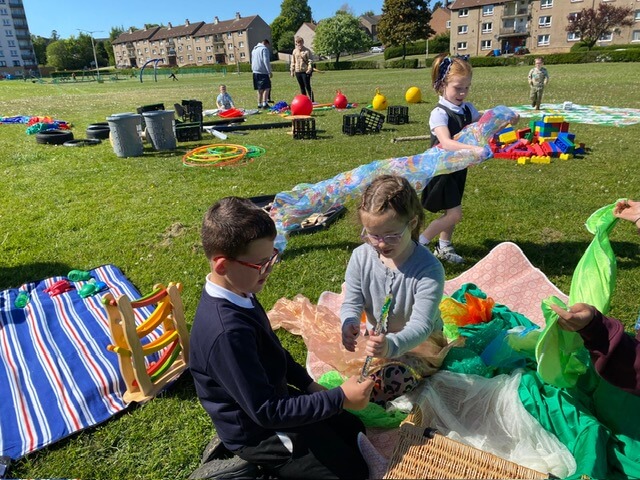 Children sitting on a blanket with colorful fabric pieces and outdoor play equipment nearby.