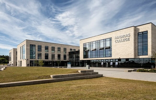 modern, multi-story Madras College building under a blue sky with light clouds. The building features a light brick facade, large glass windows, and the words 