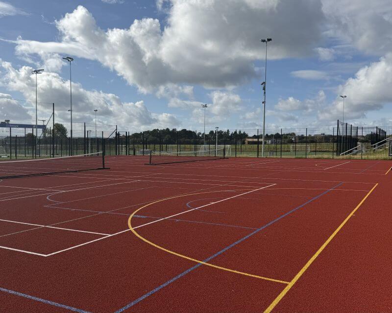 Side view of a red ash area with basketball courts painted amongst netball linings with tennis nets in the middle of the pitches