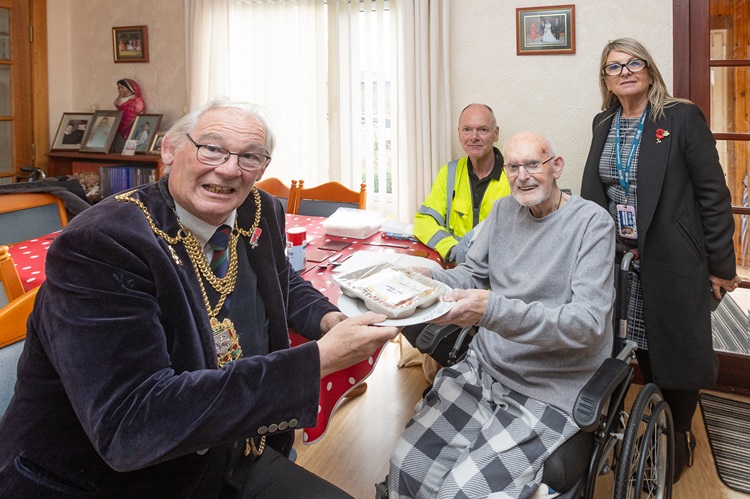 Mr Charles Middleton, in a wheelchair, receives a meal tray from Provost of Fife Jim Leishman. The Provost is wearing his ceremonial gold chain. They are indoors in a dining area with a table covered in a red cloth, chairs, and framed pictures on the wall. Two other people, Meals on Wheels driver Colin Wotherspoon and Team Manager Stella Stewart, stand nearby. Light streams in through a window with white vertical blinds in the background. - Fife celebrates Meals on Wheels Week 2025 with VIP delivery from Provost Jim Leishman