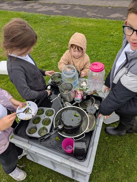 Children gathered around a mud kitchen table with pots, pans, and water containers.
