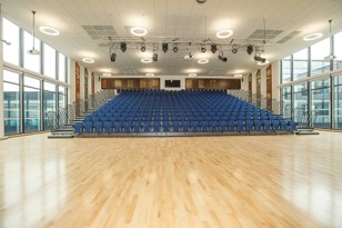 A spacious, bright assembly hall with a polished light-wood floor in the foreground. In the background, rows of blue tiered seating rise toward the back wall. Large floor-to-ceiling windows line both side walls, and overhead lighting rigs with spotlights are suspended from the high ceiling.