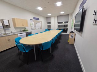 A modern meeting or staff room featuring a long, oval-shaped light wood conference table surrounded by bright blue chairs. The room has dark carpeting, a small kitchenette area with wooden cabinets on the left wall, and a large flat-screen display mounted on the right wall. Two windows with gray blinds are at the far end.
