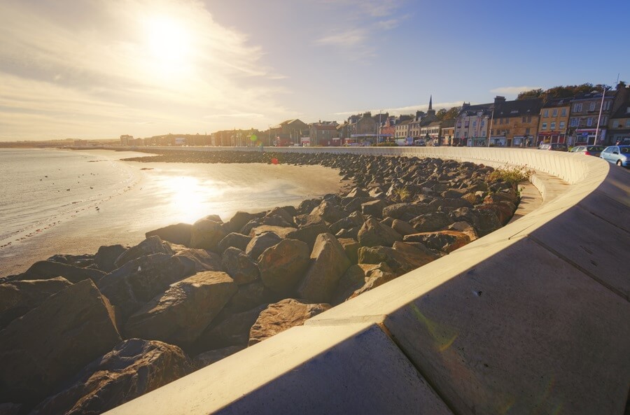 Kirkcaldy seaside promenade with a curved seawall, rocky shoreline, and buildings along the coast at sunset. - Funding boost for Kirkcaldy area projects