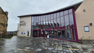 Entrance to Inverkeithing Civic Centre, a modern building with a large glass front and red framing, with a paved area leading to the entrance.