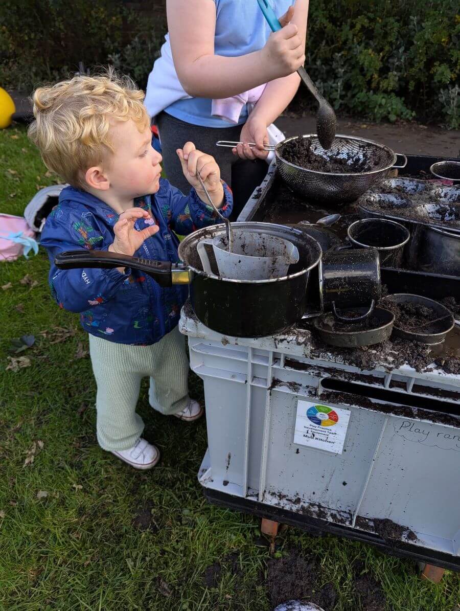 Child playing at a mud kitchen with pots and utensils on a plastic table.