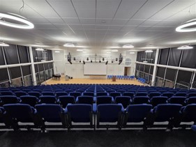An elevated view from the back of the assembly hall, looking down over rows of empty blue upholstered seats toward the stage. The stage has a light-colored floor, a large projector screen on the back wall, a podium, and a row of chairs. The large side windows are covered by dark blinds, and circular light fixtures illuminate the ceiling.