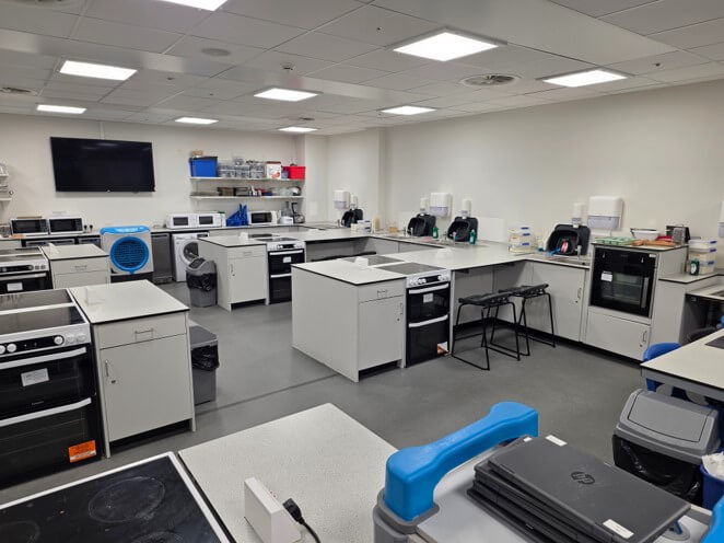 A large, modern home economics teaching kitchen filled with multiple individual white cooking stations. Each station is equipped with an electric stove, oven, counter space, and storage cabinets. In the background, there are wall-mounted microwaves, sinks, a washing machine, a flat-screen television, and various kitchen appliances.