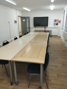 maxwell centre multi-purpose room a meeting room with a long, light-colored table surrounded by black chairs. A large television screen is mounted on the white wall at one end of the table. The floor is covered with light brown tiles. There is a doorway visible on the left side of the room and a window with blinds on the right.