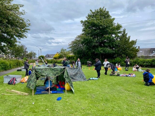 Group of people and children around a camo tarp shelter with outdoor play items scattered on grass.