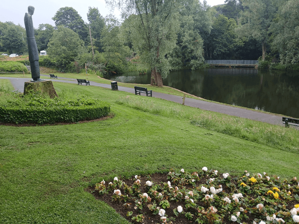 Scenic view of Riverside Park with a standing figure statue, benches, flower bed, and pond surrounded by trees