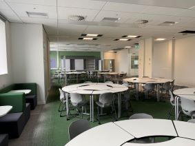 A bright, modern indoor workspace with flexible seating. Several round white tables are arranged across green carpeted flooring, each surrounded by lightweight white chairs on wheels. On the left side, low green upholstered seating lines the wall beneath a window. The ceiling has recessed lighting panels, and the room extends into another open area in the background with more tables and chairs, creating a spacious, adaptable environment designed for group work, meetings, or training.