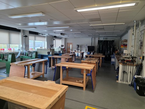A spacious wood workshop and technical classroom featuring multiple heavy wooden workbenches, each equipped with integrated vices. Larger machinery, including band saws and drill presses, line the back and side walls. The room is brightly lit by overhead fluorescent panel lighting, with a series of large windows along the left side.