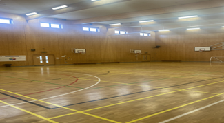 an indoor gymnasium with a polished wooden floor marked with colorful lines for various sports. Basketball hoops are visible on the wooden walls. There are also double doors on the left side of the image.