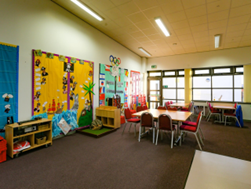 a creche room with a dark carpet. There are colourful posters on the walls, and tables with red chairs are arranged in the room.