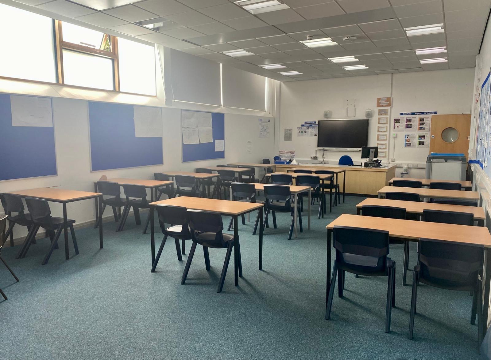Empty classroom with individual desks and chairs, a teaching desk and screen at the front, and display boards on the walls