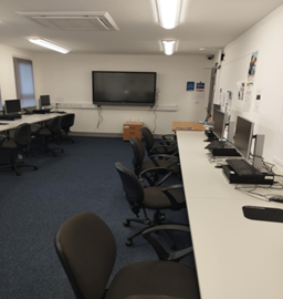 a computer lab with rows of white desks and black chairs. Computer monitors, keyboards, and mice are set up on the desks. A large flat-screen television is mounted on the wall at the back of the room. The floor is covered with a dark blue carpet. There are posters and notices on the white walls. a computer lab with rows of white desks and black chairs. Computer monitors, keyboards, and mice are set up on the desks. A large flat-screen television is mounted on the wall at the back of the room. The floor is covered with a dark blue carpet. There are posters and notices on the white walls.
