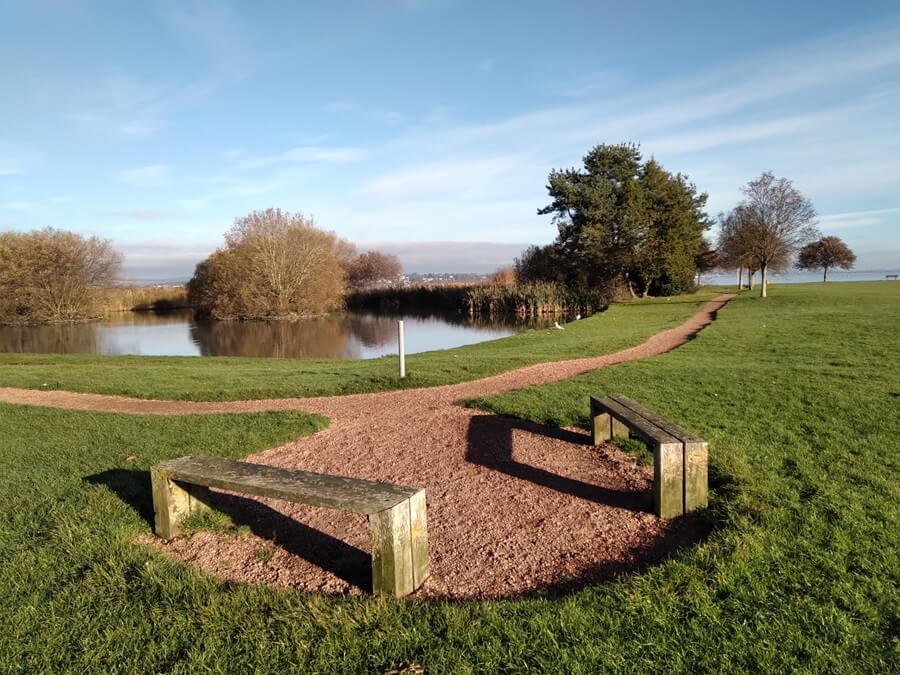 Peaceful outdoor scene with two wooden benches on a gravel path that splits toward a small lake. Trees, bushes, and green grass surround the area under a partly cloudy sky