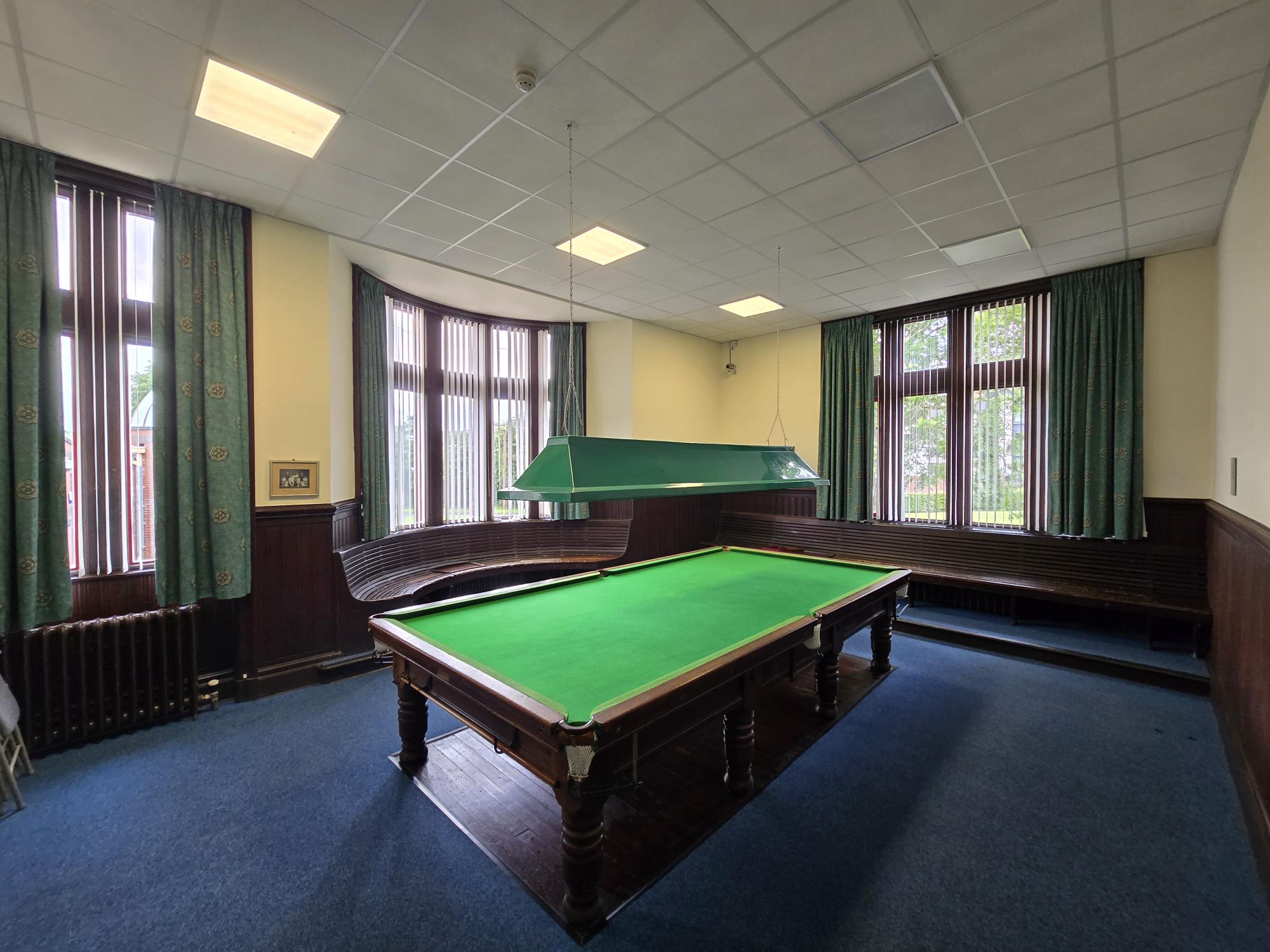 Room with a full-size snooker table, green overhead light, dark wood seating around the walls, and large curtained windows