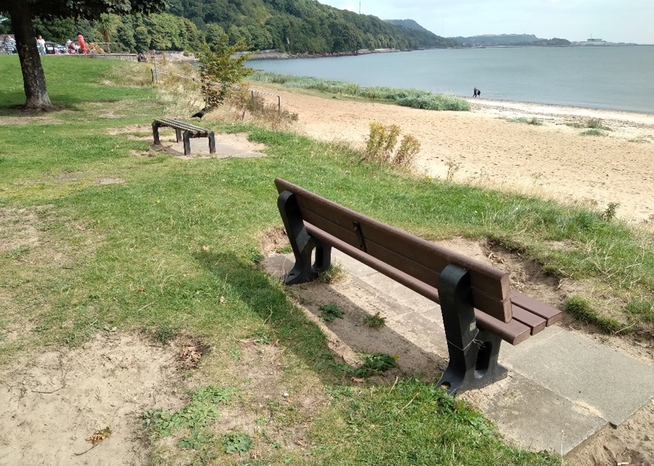 Two benches on grass near a beach with water, trees, and hills in the background
