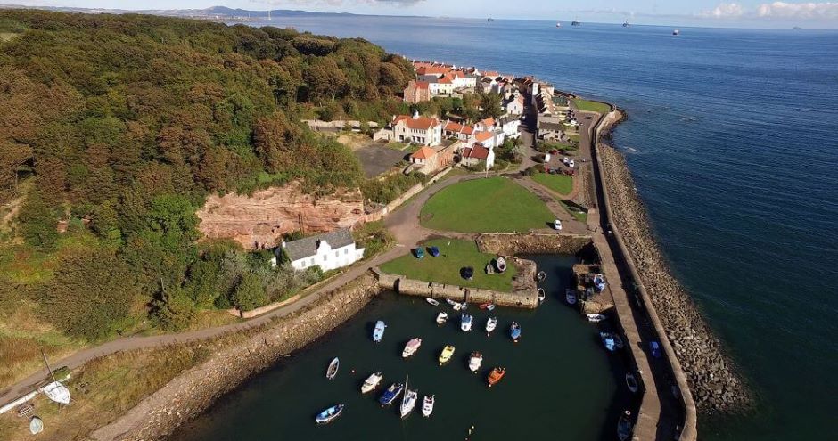 Aerial view of West Wemyss village on the Fife coast, showing a small harbour with moored boats in calm water, a curving sea wall along the shoreline, and rows of red-roofed cottages clustered beside a green space. Behind the houses, a wooded hillside rises with exposed rock faces, while the open sea stretches into the distance under clear skies.