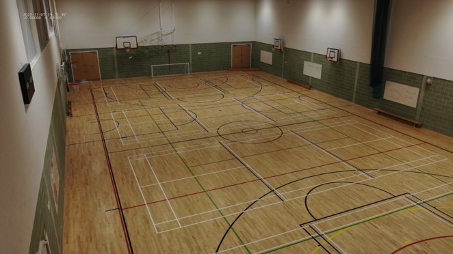 A high-angle view of a large indoor sports hall with a polished wooden floor, marked with various coloured lines for different sports. Basketball hoops and a soccer goal are visible along the walls, which feature green brickwork on the lower half and white paint on the upper half.
