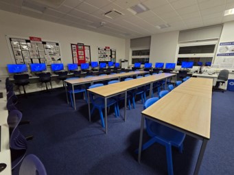 A school IT suite featuring rows of desktop computers with bright blue screens arranged along the back and side walls. Several long, light-wood student desks with blue chairs sit in the center of the room. The room has dark blue carpeting, white walls with educational posters, and windows at the far end.