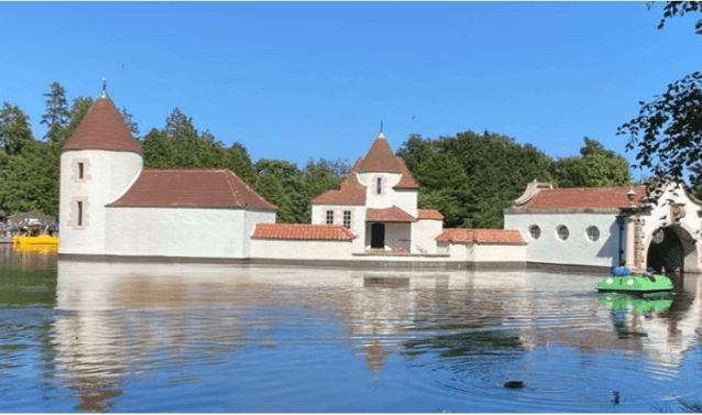 A white, castle-like structure with orange tiled roofs sits in the middle of a calm lake under a clear blue sky