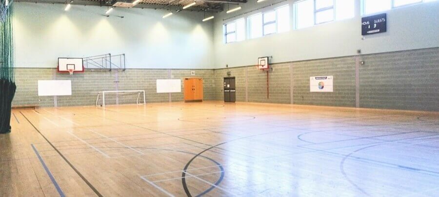games hall An indoor sports hall. The wooden floor has court lines. Basketball hoops and a goal stand.