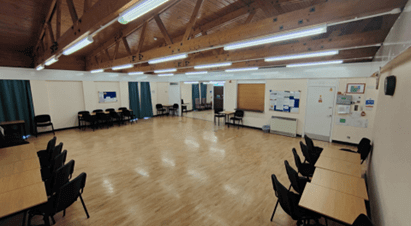 A wide shot of an empty community hall with a polished wooden floor, tables and chairs arranged on the sides, and a mirrored wall at the far end.