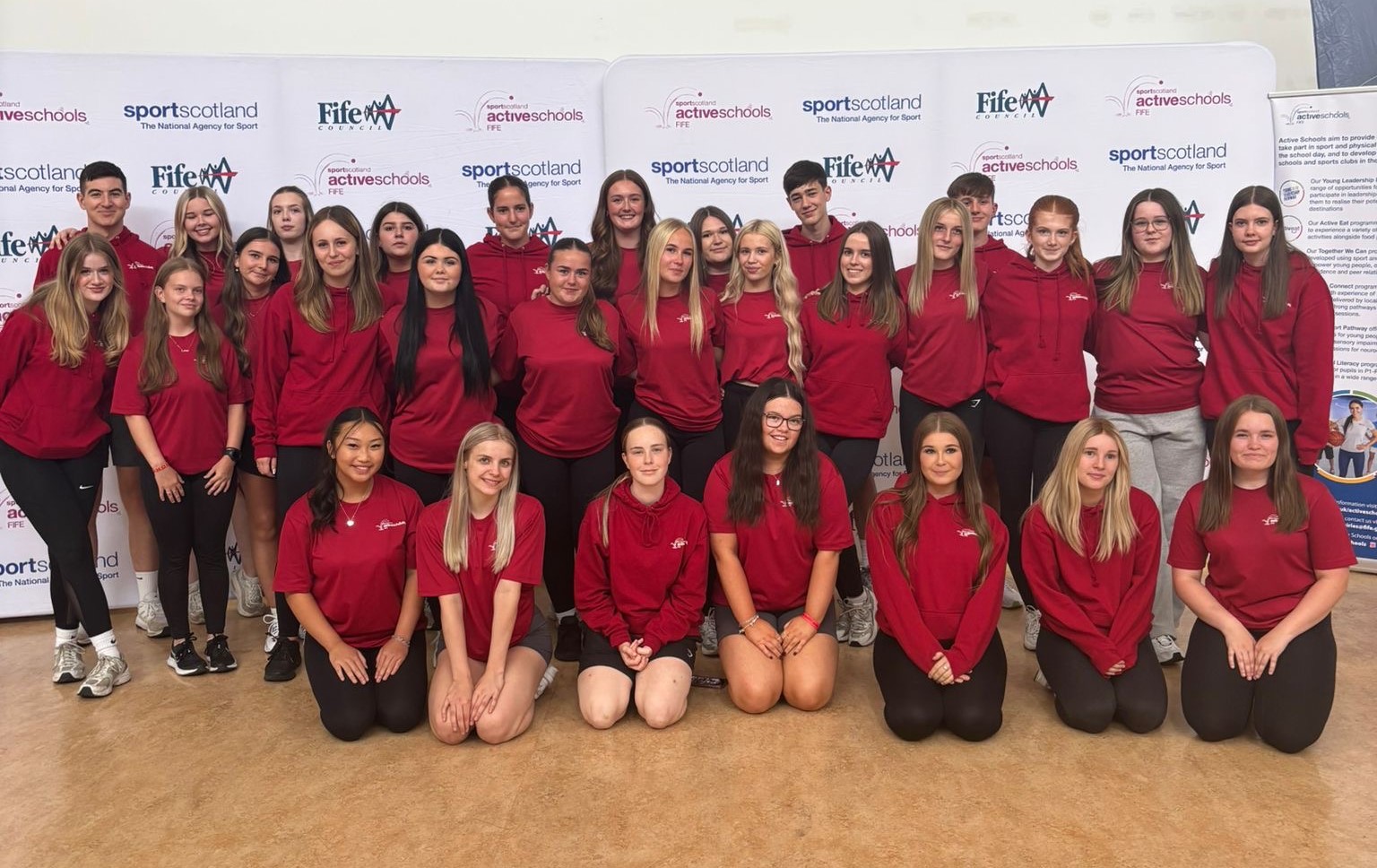 Active Schools Platinum ambassadors - A group of young people wearing matching red shirts and hoodies pose together indoors in front of banners for Fife Council, SportScotland, and Active Schools. - Fife Active Schools celebrates inspiring Young Ambassador Day 2025