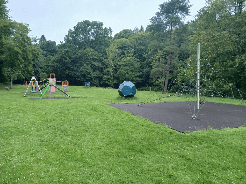 Playground with slides and climbing structures in a grassy park area surrounded by trees