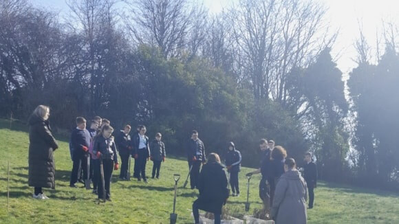 A group of children and adults stand on a grassy slope in Cowdenbeath Public Park, gathered around planting tools and saplings, with trees in the background on a bright day. - Enhancing biodiversity at Cowdenbeath Public Park