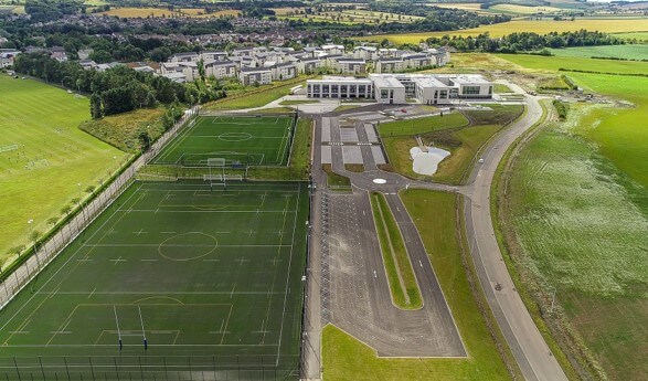 An aerial view of the Madras College campus, showing two large green artificial turf (astro) sports pitches in the foreground. Beside the pitches is a large paved parking lot and a drop-off loop. In the background sits the modern school building, flanked by green countryside and a residential neighborhood.