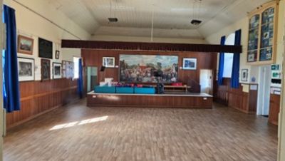 Main hall of a community building with a large open wooden floor and a raised stage at the far end. The stage has a painted mural backdrop and tables set up, while framed pictures and blue curtains line the wood-panelled walls.
