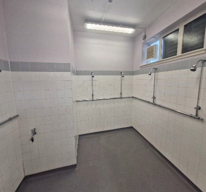 A shower room. White tiles line the walls. Multiple shower heads are on the wall. A shower room. White tiles line the walls. Multiple shower heads are on the wall.