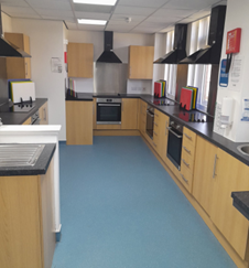 a kitchen classroom with light wood cabinets, black countertops, and stainless steel sinks. Black ventilation hoods are above the ovens and stovetops. The floor is blue. a kitchen classroom with light wood cabinets, black countertops, and stainless steel sinks. Black ventilation hoods are above the ovens and stovetops. The floor is blue.