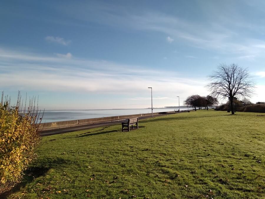 Coastal park with grass and a bench facing the River Tay. A path runs beside the water, lined with trees under a lightly clouded sky