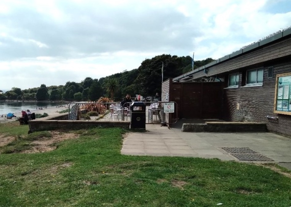 Beachside building with outdoor seating, a paved walkway, and people in the background near trees and the shore.