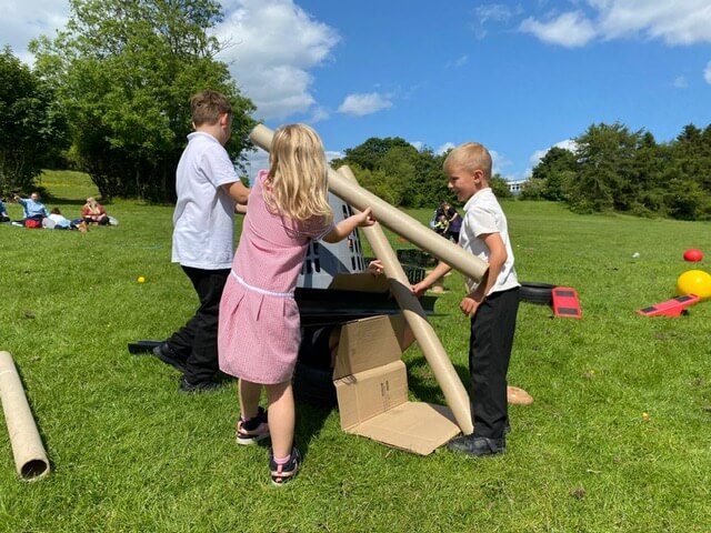 Three children building a structure with cardboard and large cardboard tubes on grass.