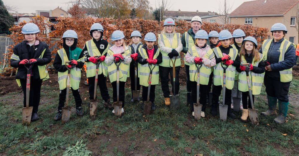 Pupils and teachers standing outdoors on a grassy area, wearing safety vests, hard hats, and gloves. Each person is holding a shovel.