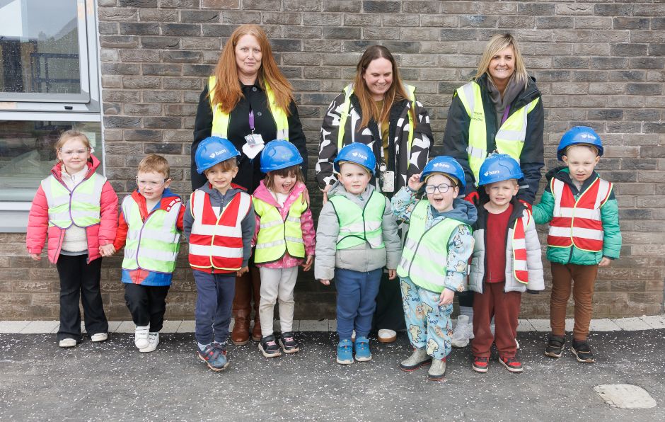 A group of young children and three adults stand in front of a brick wall. The children are wearing high visibility vests, with some also wearing blue hard hats. The adults behind them are also in high visibility clothing.