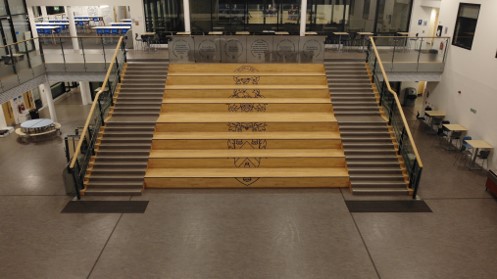 A wide indoor atrium featuring a large, symmetrical central staircase leading up to a mezzanine level. The middle section of the stairs consists of wide, light-wood seating steps adorned with a vertical line of graphic school crests. Functional gray-carpeted steps with handrails flank both sides of the wooden seating. The surrounding area has open corridors and scattered student tables.