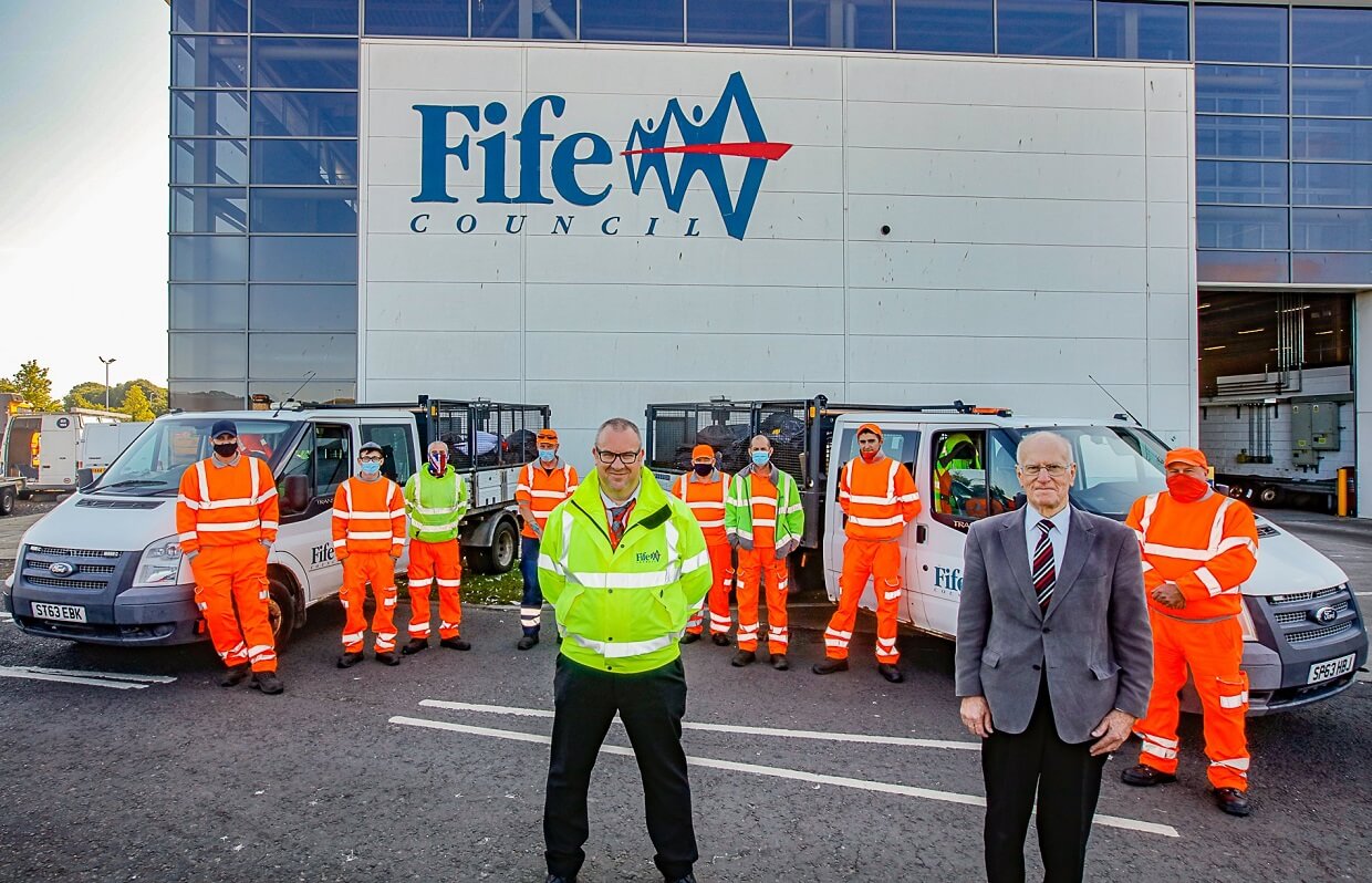 Group photo of Fife Council workers in high-visibility orange uniforms standing in front of council vehicles, with two officials in the foreground. The Fife Council logo is displayed on the building in the background.
