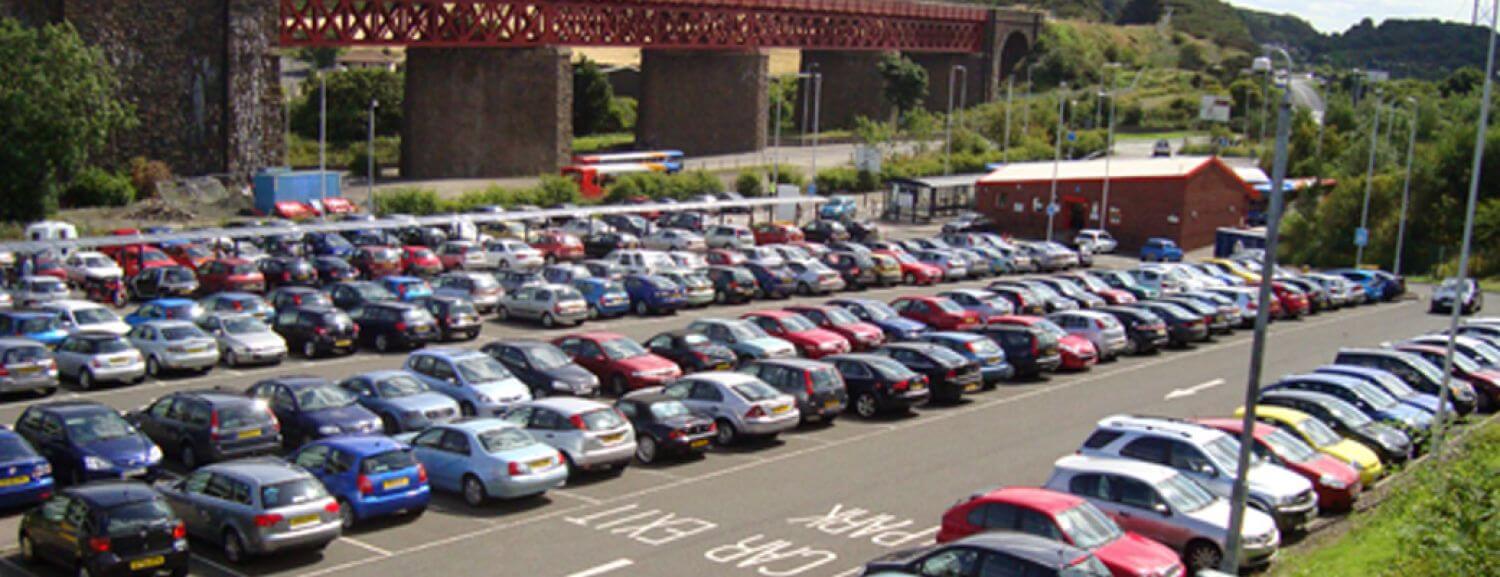 Ferrytoll Park & Ride with rows of parked cars, a red bridge supported by stone pillars in the background, nearby buildings and greenery, and “CAR PARK EXIT” painted on the ground.